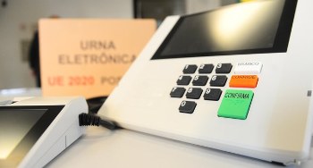 Urna Eletrônica em primeiro plano e uma caixa de papelão ao fundo desfocada, com a escrita "Urna...