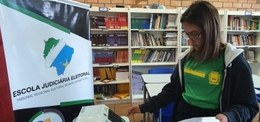 A imagem apresenta uma menina de camiseta verde pressionando uma urna eletrônica em uma sala com...