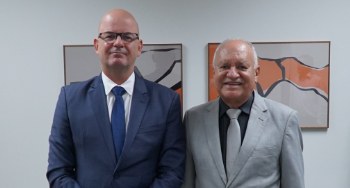 Foto com dois homens vestidos de terno, sorrindo para a foto, os dois de meia idade, um deles us...