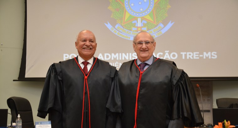 Imagem de foto oficial com dois homens vestidos com becas para posse solene sorrindo para a foto...