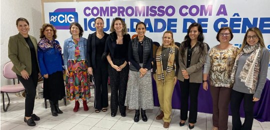 Foto de um grupo de mulheres alinhadas em fileira sorrindo para a foto à frente de um painel com...