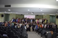 Foto em auditório com dezenas de pessoas organizadas em fileiras no palco de frente para cadeira...